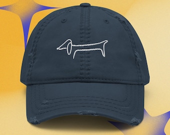 Puede incluir: Una gorra de béisbol azul marino desgastada con un gráfico bordado en hilo blanco de un perro salchicha centrado en el panel frontal. La gorra tiene una visera curva con bordes deshilachados y una corona estructurada con botón superior.