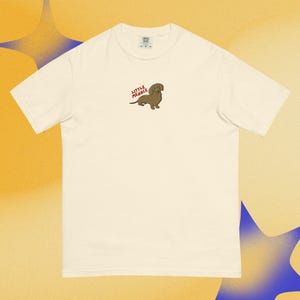 Puede incluir: Una camiseta de manga corta de color crema con un pequeño gráfico bordado en la parte superior del pecho. El gráfico muestra un perro salchicha marrón con el texto rojo "LITTLE MENACE" colocado sobre el lomo del perro en una fuente sencilla.