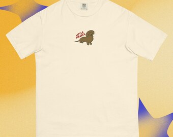 Puede incluir: Una camiseta de manga corta de color crema con un pequeño gráfico bordado en la parte superior del pecho. El gráfico muestra un perro salchicha marrón con el texto rojo "LITTLE MENACE" colocado sobre el lomo del perro en una fuente sencilla.