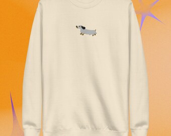 Puede incluir: Una sudadera de cuello redondo color crema con un pequeño bordado centrado de un perro salchicha gris claro con orejas y patas marrones. El jersey de manga larga presenta cuello, puños y dobladillo acanalados para un acabado sencillo.