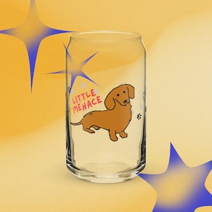 Puede incluir: Un vaso transparente con forma de lata que presenta una ilustración de un perro salchicha marrón y el texto rojo "LITTLE MENACE" impreso en el frente. El diseño incluye pequeñas huellas negras y contornos de huesos alrededor.