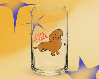 Puede incluir: Un vaso transparente con forma de lata que presenta una ilustración de un perro salchicha marrón y el texto rojo "LITTLE MENACE" impreso en el frente. El diseño incluye pequeñas huellas negras y contornos de huesos alrededor.