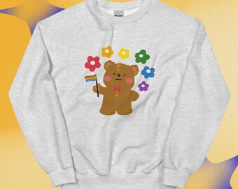 Puede incluir: Una sudadera gris claro de cuello redondo con un gráfico de un oso marrón. El oso tiene mejillas rosadas, una pajarita roja y una fila de botones arcoíris. Sostiene una bandera arcoíris, rodeado por un semicírculo de flores rojas, amarillas, verdes, azules y moradas.