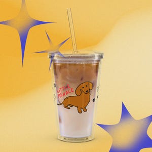Puede incluir: Un vaso de plástico transparente con tapa y pajita, lleno de una bebida de café helado. El exterior presenta una ilustración de un perro salchicha marrón y el texto rojo "LITTLE MENACE", junto a pequeñas huellas negras y huesos blancos.