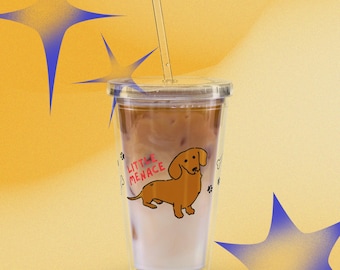 Puede incluir: Un vaso de plástico transparente con tapa y pajita, lleno de una bebida de café helado. El exterior presenta una ilustración de un perro salchicha marrón y el texto rojo "LITTLE MENACE", junto a pequeñas huellas negras y huesos blancos.