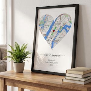 May include: A framed print featuring a heart-shaped map of New York City with a red heart icon over Central Park. Below the map, the text reads: Tomi and Jasmine, Engaged, Central Park, NYC, 6.6.26. The print is in a black frame.