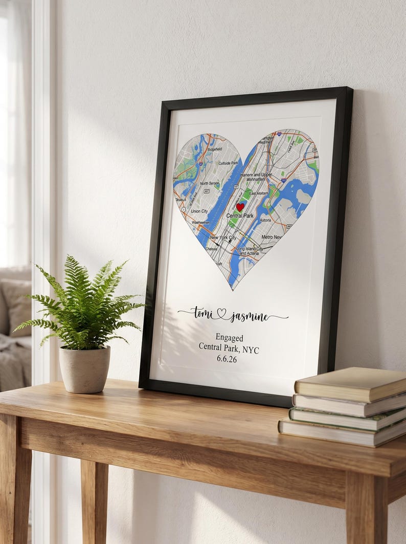 May include: A framed print featuring a heart-shaped map of New York City with a red heart icon over Central Park. Below the map, the text reads: Tomi and Jasmine, Engaged, Central Park, NYC, 6.6.26. The print is in a black frame.