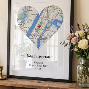 May include: A framed wall art print featuring a heart-shaped map of New York City with a red heart icon over Central Park. Below the map, the text reads: Tomi and Jasmine, Engaged, Central Park, NYC, 6.6.26, in black script and serif fonts.