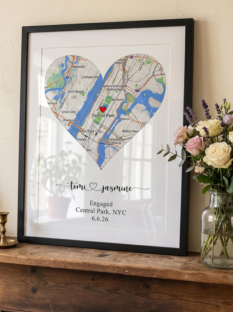 May include: A framed wall art print featuring a heart-shaped map of New York City with a red heart icon over Central Park. Below the map, the text reads: Tomi and Jasmine, Engaged, Central Park, NYC, 6.6.26, in black script and serif fonts.