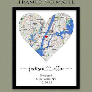 May include: A framed map of New York City shaped like a heart with a red heart icon over Manhattan. Below the map, the text reads: jackson ♡ ellie, Engaged, New York, NY, 12.24.25. The top of the image includes the text FRAMED NO MATTE.