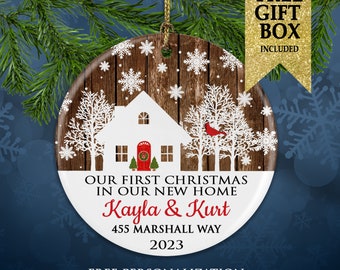 May include: A round Christmas ornament with a house design, snowflakes, and a red cardinal. The ornament has the text "OUR FIRST CHRISTMAS IN OUR NEW HOME" and personalized names and address. A gold gift box is included.