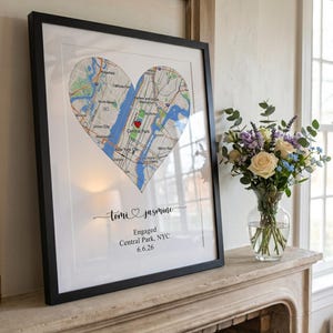May include: A framed wall art print featuring a heart-shaped map of New York City with a red heart icon over Central Park. Below the map, the text reads: Tomi & Jasmine, Engaged, Central Park, NYC, 6.6.26. The print is in a black frame.