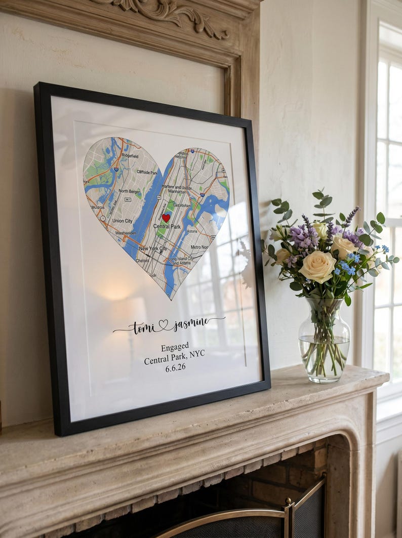 May include: A framed wall art print featuring a heart-shaped map of New York City with a red heart icon over Central Park. Below the map, the text reads: Tomi & Jasmine, Engaged, Central Park, NYC, 6.6.26. The print is in a black frame.