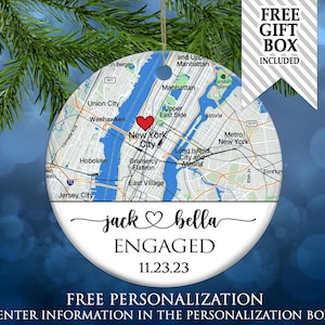 May include: A round ceramic ornament featuring a map of New York City with a red heart icon. Below the map, the text reads jack, a heart symbol, bella, ENGAGED, and 11.23.23 in black script and serif fonts. A gold string is attached at the top.