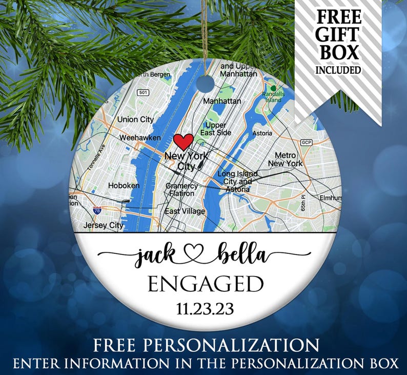 May include: A round ceramic ornament featuring a map of New York City with a red heart icon. Below the map, the text reads jack, a heart symbol, bella, ENGAGED, and 11.23.23 in black script and serif fonts. A gold string is attached at the top.