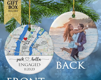 May include: A round ceramic ornament featuring a map of New York City with a red heart icon on the front, along with the text jack & bella ENGAGED 11.23.23. The back displays a photograph of a couple embracing on a beach at sunset.
