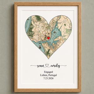 May include: A framed map of Lisbon, Portugal, shaped as a heart with a red heart icon marking the city. Below the map, the text reads: sean heart emily, Engaged, Lisbon, Portugal, 7.23.2026. The print is housed in a light wood frame with a white mat.