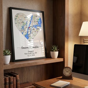 May include: A framed wall art print featuring a heart-shaped map of New York City with a red heart icon marking a specific location. Below the map, the text reads: lauren and austin, Engaged, New York City, NY, 12.31.2024 in black script and print.