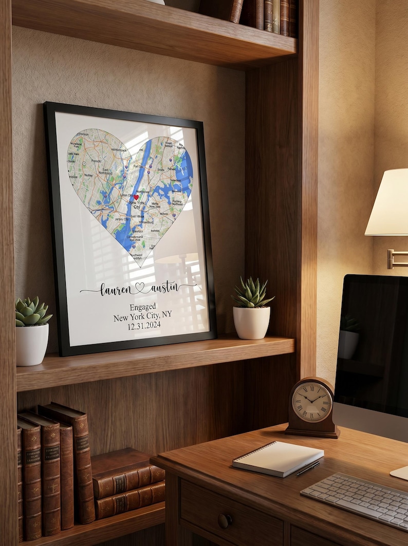 May include: A framed wall art print featuring a heart-shaped map of New York City with a red heart icon marking a specific location. Below the map, the text reads: lauren and austin, Engaged, New York City, NY, 12.31.2024 in black script and print.