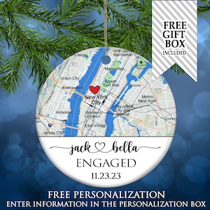 May include: A round ceramic ornament featuring a map of New York City with a red heart icon over Manhattan. Below the map, the text reads jack heart symbol bella, ENGAGED, and 11.23.23. The ornament has a gold hanging string at the top.