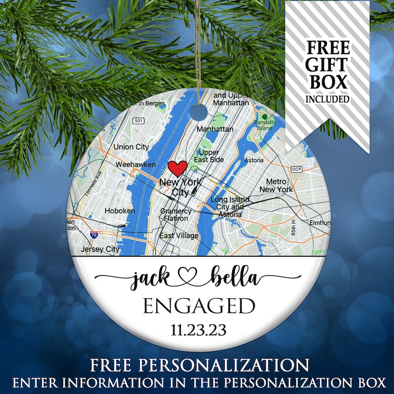 May include: A round ceramic ornament featuring a map of New York City with a red heart icon over Manhattan. Below the map, the text reads jack heart symbol bella, ENGAGED, and 11.23.23. The ornament has a gold hanging string at the top.