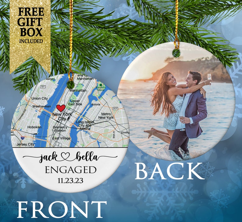 May include: A round ceramic ornament featuring a map of New York City with a red heart icon on the front, along with the text "jack & bella ENGAGED 11.23.23". The back displays a photograph of a couple embracing in the ocean at sunset.