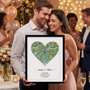 May include: A framed rectangular wall art piece featuring a heart-shaped map of Varenna, Italy, with a red heart icon marking the location. Below the map, the text reads: nina and dan, Engaged, Varenna, Italy, 5.19.2026, in black lettering.