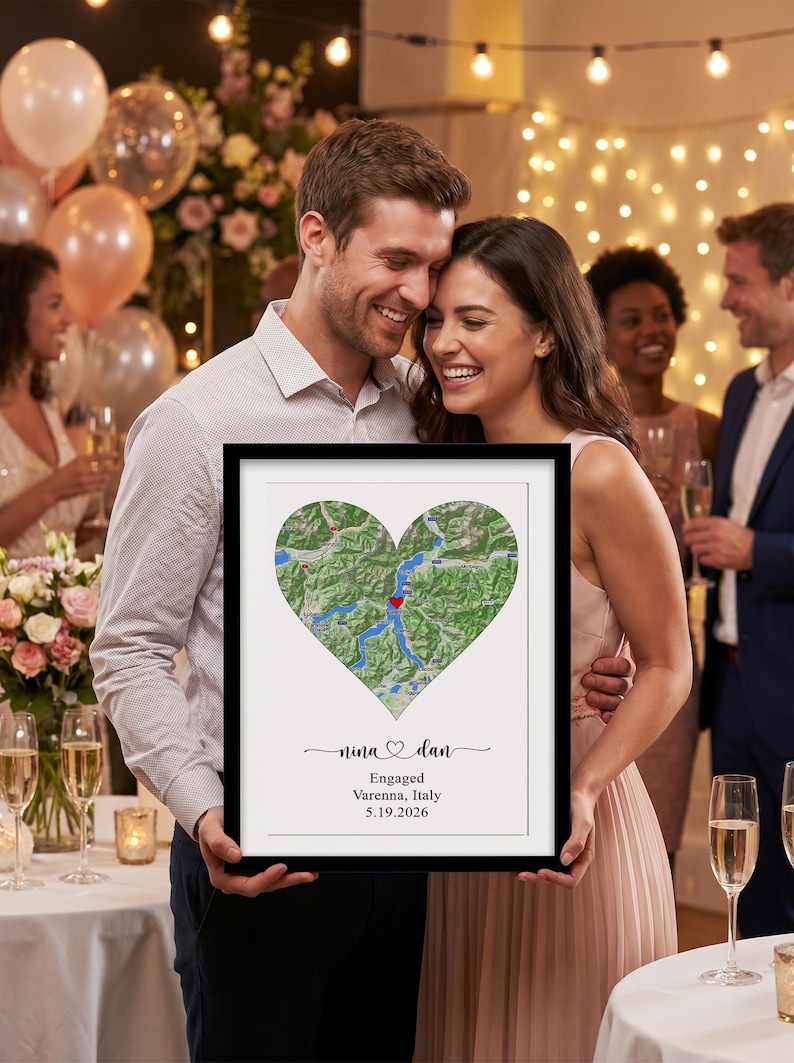 May include: A framed rectangular wall art piece featuring a heart-shaped map of Varenna, Italy, with a red heart icon marking the location. Below the map, the text reads: nina and dan, Engaged, Varenna, Italy, 5.19.2026, in black lettering.