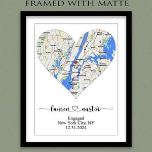 May include: A framed print with a white matte featuring a heart-shaped map of New York City with a red heart icon. Below the map, the text reads: lauren ♡ austin, Engaged, New York City, NY, 12.31.2024. The frame is black and rectangular.