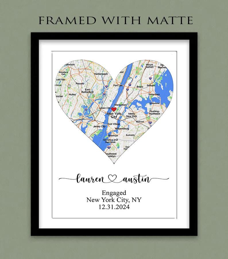 May include: A framed print with a white matte featuring a heart-shaped map of New York City with a red heart icon. Below the map, the text reads: lauren ♡ austin, Engaged, New York City, NY, 12.31.2024. The frame is black and rectangular.