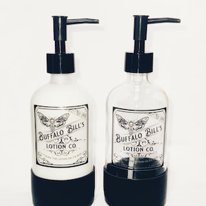 May include: Two glass bottles with black pump tops and black bases. The bottles are filled with white lotion and have black and white labels that read "Buffalo Bill's Lotion Co. Est. 1999" with a moth graphic.