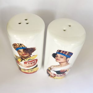 May include: A pair of white ceramic salt and pepper shakers. Each shaker features a portrait of a woman wearing a hat and jewelry. The shakers have three holes on top for dispensing.