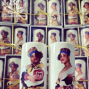 May include: White ceramic candle holders featuring a colorful portrait of a woman wearing a hat and jacket. The candle holders are packaged in individual boxes. The text "REIMAGINED EMPIRE" is visible in the background.
