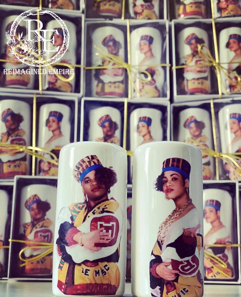 May include: White ceramic candle holders featuring a colorful portrait of a woman wearing a hat and jacket. The candle holders are packaged in individual boxes. The text "REIMAGINED EMPIRE" is visible in the background.