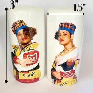 May include: A pair of white ceramic salt and pepper shakers, each 3 inches tall and 1.5 inches wide, featuring a colorful portrait of a woman wearing a hat and gold jewelry. The shakers have a retro design with the text "LE MSC MAGIC".