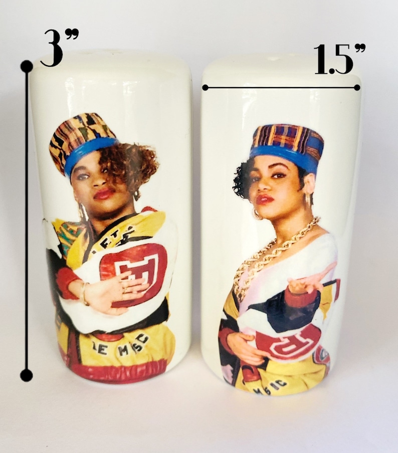 May include: A pair of white ceramic salt and pepper shakers, each 3 inches tall and 1.5 inches wide, featuring a colorful portrait of a woman wearing a hat and gold jewelry. The shakers have a retro design with the text "LE MSC MAGIC".