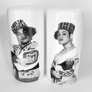 May include: Two white ceramic salt and pepper shakers. Each shaker features a black and white image of a woman wearing a hat and a jersey with the letters "E.T.S." and "M.S.C." on the bottom. The shakers are cylindrical.