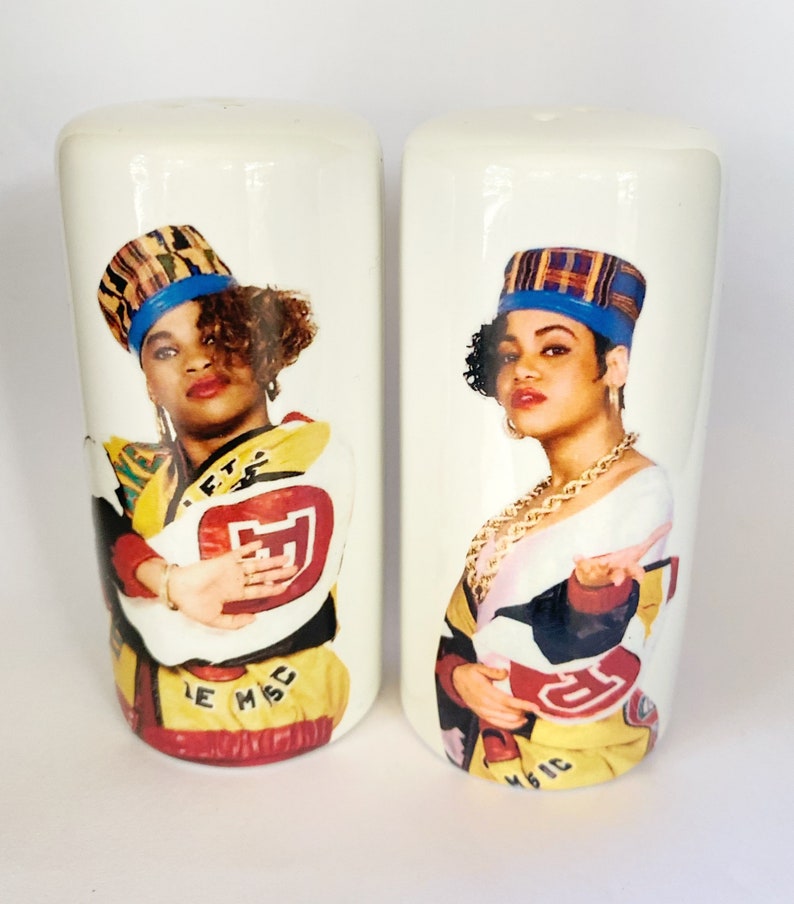 May include: A pair of white ceramic salt and pepper shakers. Each shaker features a colorful illustration of a woman wearing a hat and jacket. The jackets have the text "LE MSC MAGIC".