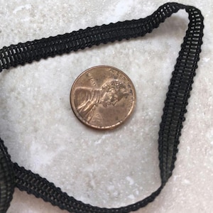 May include: Black decorative trim with a woven design. A penny is next to the trim for size comparison. The trim is likely used for sewing, crafting, or adding detail to clothing or accessories.
