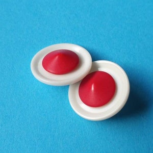 May include: Two circular buttons featuring a smooth white outer rim and a raised, conical red center. The buttons have a glossy finish and are positioned slightly overlapping each other, showcasing their distinct two-tone color scheme and shape.