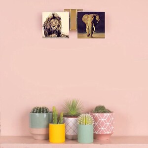 May include: Two framed prints of animal photographs. The top print shows a lion with a brown mane. The bottom print shows an elephant with large ears and tusks. The prints are hanging on a light pink wall above a shelf with five potted succulents.