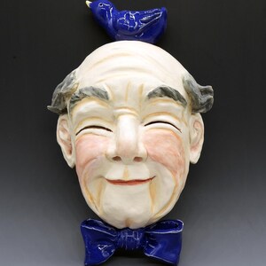 May include: A ceramic wall mask depicting a smiling face with closed eyes, rosy cheeks, and a blue bow tie. A small blue bird sits atop the head. The mask is painted in shades of cream, pink, and blue.