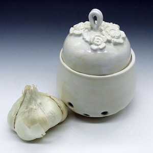 May include: A cream-colored ceramic garlic keeper with a floral design on the lid. The keeper has a round body with three small holes and a matching lid with a loop handle. A whole garlic bulb sits beside the keeper.