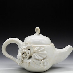 May include: A white ceramic teapot with a floral design. The teapot features a sculpted rose and other flowers on the side. It has a curved handle and a spout. The lid has a loop handle.