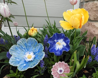 May include: A collection of ceramic flower decorations in a garden setting. The flowers are blue, pink, and yellow with white accents. The flowers are arranged in a row on a bed of green plants.