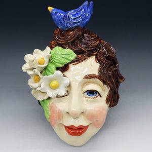May include: A ceramic face sculpture with brown hair, blue eyes, and red lips. A blue bird sits atop the head, and white flowers with yellow centers adorn one side. The face has rosy cheeks and a winking expression.