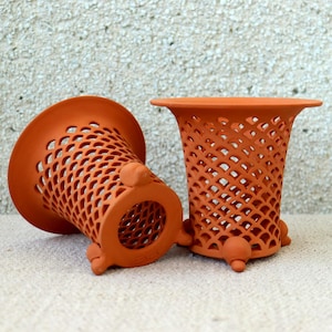 Può includere: Due vasi traforati color terracotta. Un vaso è in posizione verticale, l'altro è di lato. Ognuno ha una parte superiore svasata, un corpo a forma di reticolo e tre piedini. I vasi sono fatti di argilla marrone rossiccia e hanno un aspetto rustico.