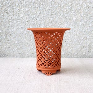 Può includere: Un vaso traforato color terracotta, con la parte superiore svasata e un motivo geometrico. Il vaso ha una forma cilindrica con un design decorativo e poggia su tre piedini. Lo sfondo è una superficie strutturata di colore chiaro.
