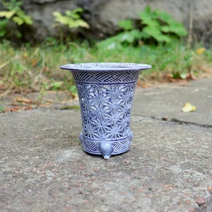 May include: A light blue ceramic vase with a wide, flared rim and a tapered body. The surface features an intricate, cut-out star pattern across the centre, bordered by geometric, cross-hatched bands at the top and bottom, resting on three feet.