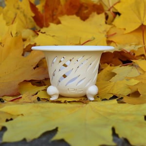 May include: A cream-colored, decorative planter with a flared rim and three small feet, set against a backdrop of vibrant yellow autumn leaves. The planter has a cut-out design. The planter is approximately 4 inches tall.